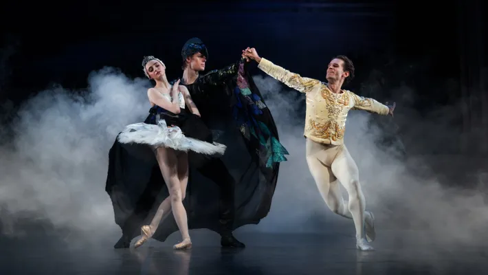 A man in a cloak embraces a ballerina: around them a male dancer dances.