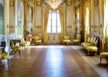 Ett rum med möbler i gult längs väggarna, stora fönster där solljuset strilar igenom.