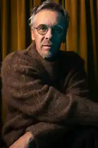 Person with glasses in brown sweater sits in front of yellow curtains, face partially hidden.