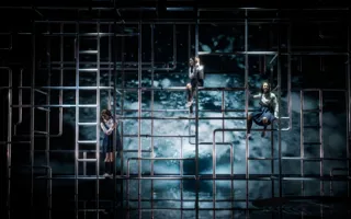 Against a water backdrop, a multitude of pipes are visible: three women are climbing the pipes.