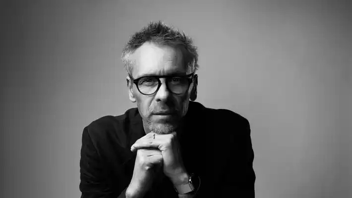 Person står med sammanlåsta händer framför sig, ansiktet suddigt och delvis dolt, mot enfärgad bakgrund.