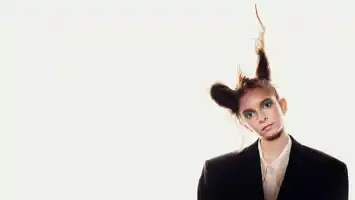 A young woman, wearing a dark jacket and with her hair tied in two buns, her head tilted slightly to the side, against a white background.