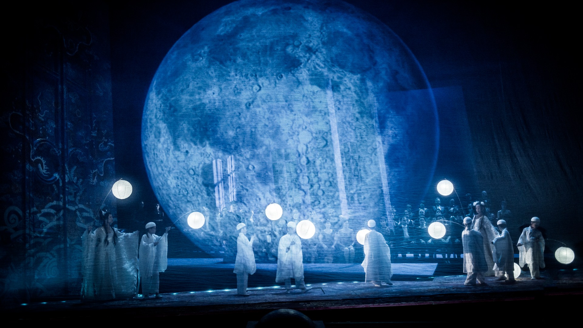 En scenbild med flera personer som rör sig i ett blåtonat landskap, i bakgrunden syns en stor himlakropp.