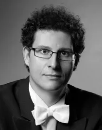 A close-up of a man with curly hair, he wears glasses and wears a tailcoat.
