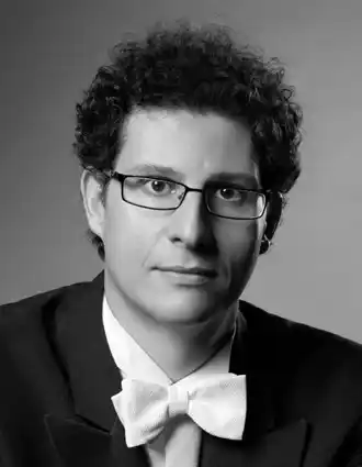 A close-up of a man with curly hair, he wears glasses and wears a tailcoat.