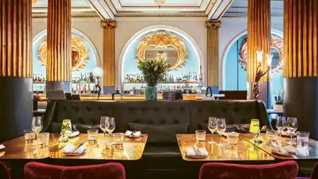 An interior with laid tables and a bar in the background, richly decorated with gold columns.