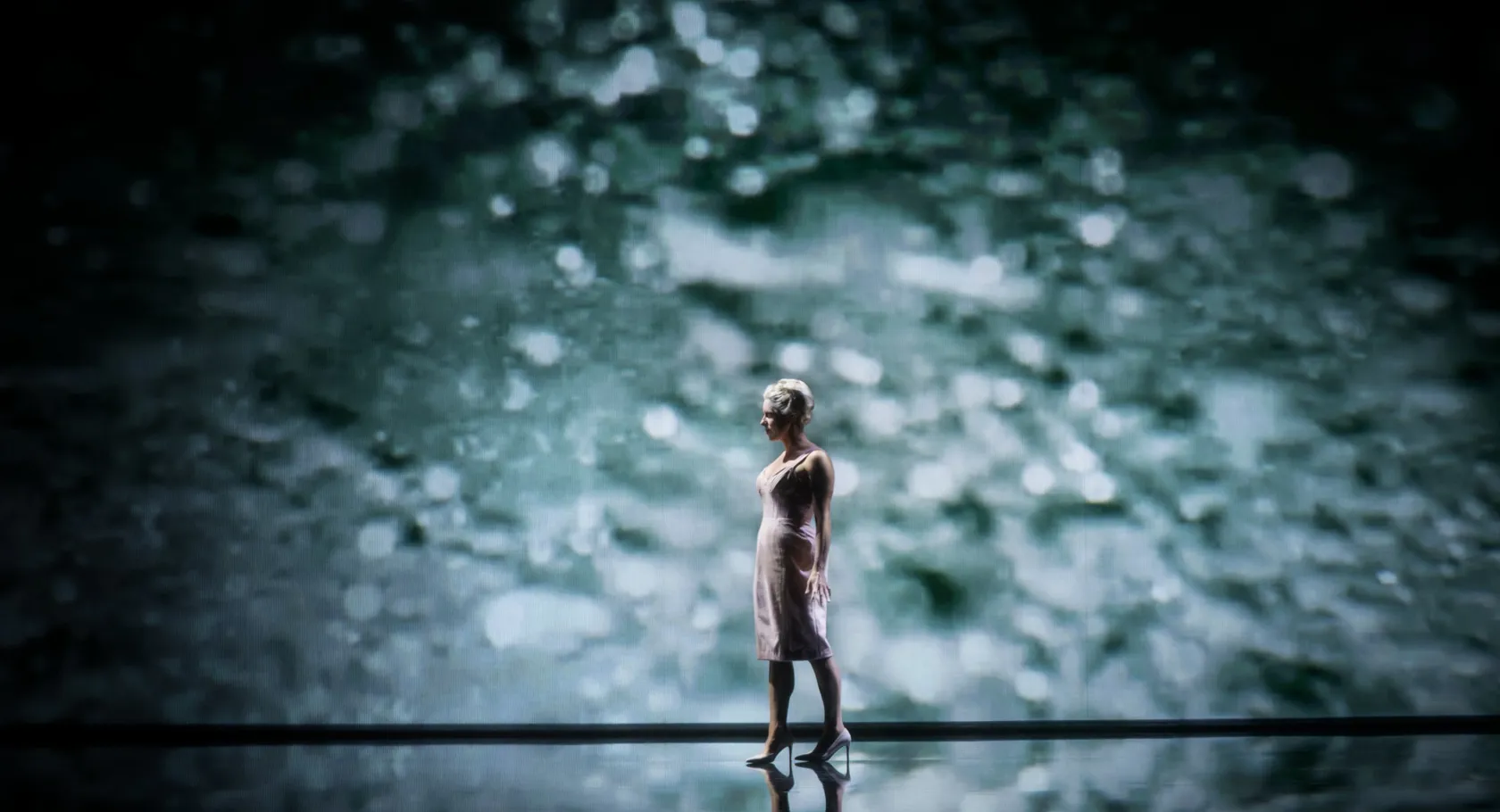 A lone woman on a stage, projected water visible in the background.