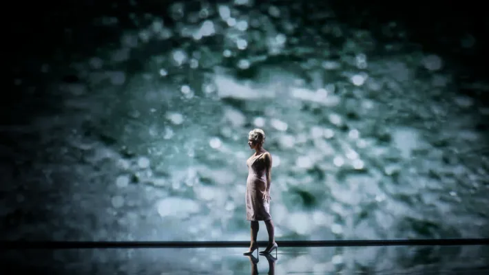 A lone woman on a stage, projected water visible in the background.