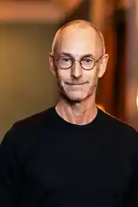 A smiling man in a black shirt and glasses against a blurred background with multiple light sources.