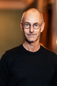 En leende man i svart tröja och glasögon mot en suddig bakgrund med flera ljuskällor.