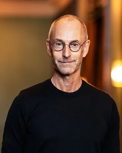 A smiling man in a black shirt and glasses against a blurred background with multiple light sources.