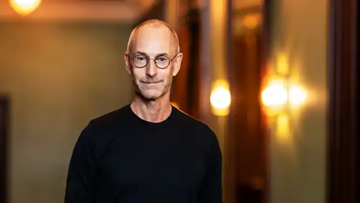 En leende man i svart tröja och glasögon mot en suddig bakgrund med flera ljuskällor.