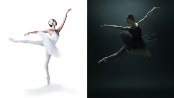 A ballerina in a white costume balances on one toe, the other leg is stretched back, opposite her a ballerina dressed in black makes a big jump.