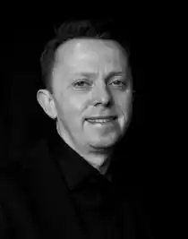 En närbild på en leende man, klädd i mörka kläder, mot en mörk bakgrund.