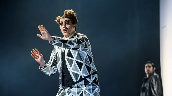 A man in a graphically patterned suit holds his hands up in front of him, another man is visible in the background.