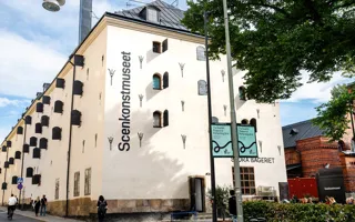 En vit byggnad med många fönsterluckor och stort träd framför.