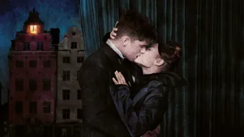 Two people kiss each other passionately in front of a dark city skyline with an illuminated window in the background.