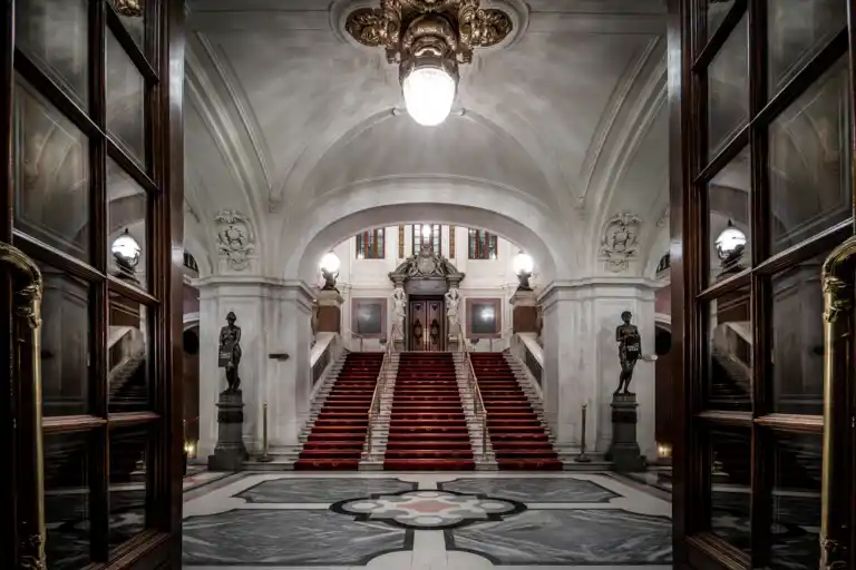 View of a large entrance hall, a staircase with red carpet leads up to a door.