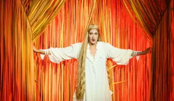 En kvinna med långt blont hår och vit klänning står framför ett orange draperi.