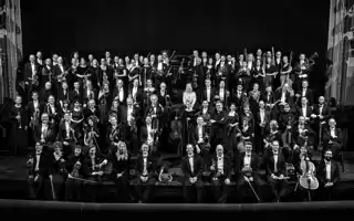 About a hundred dressed-up people stand on a stage, several carrying instruments.