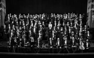 About a hundred dressed-up people stand on a stage, several carrying instruments.