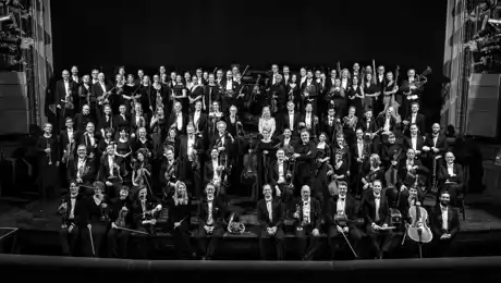About a hundred dressed-up people stand on a stage, several carrying instruments.