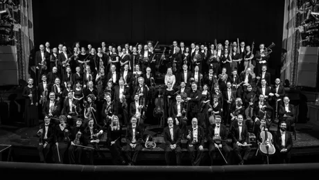 About a hundred dressed-up people stand on a stage, several carrying instruments.