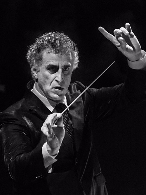 A man in a suit raising both hands, in one hand a conductor's baton.