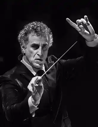 A man in a suit raising both hands, in one hand a conductor's baton.