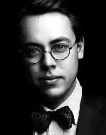 Porträttbild av en ung man med glasögon, kostym och fluga mot mörk bakgrund.