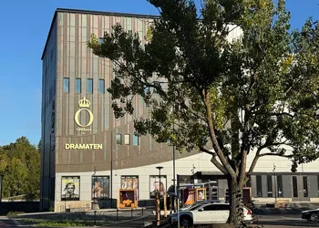 En modern byggnad ligger framför en skogsdunge, i förgrunden ett stort träd.