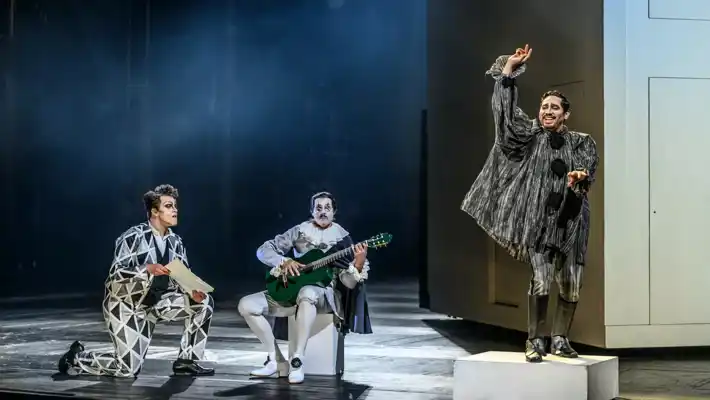 A man kneels in front of a guitarist, to their right a man stands on a cube and gestures.