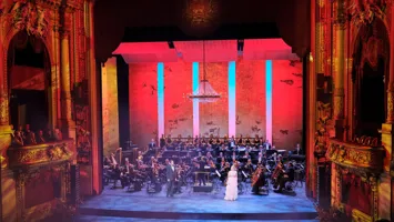 En vackert upplyst scen, omgärdad av gulddekor, med orkester och sångare i förgrunden.