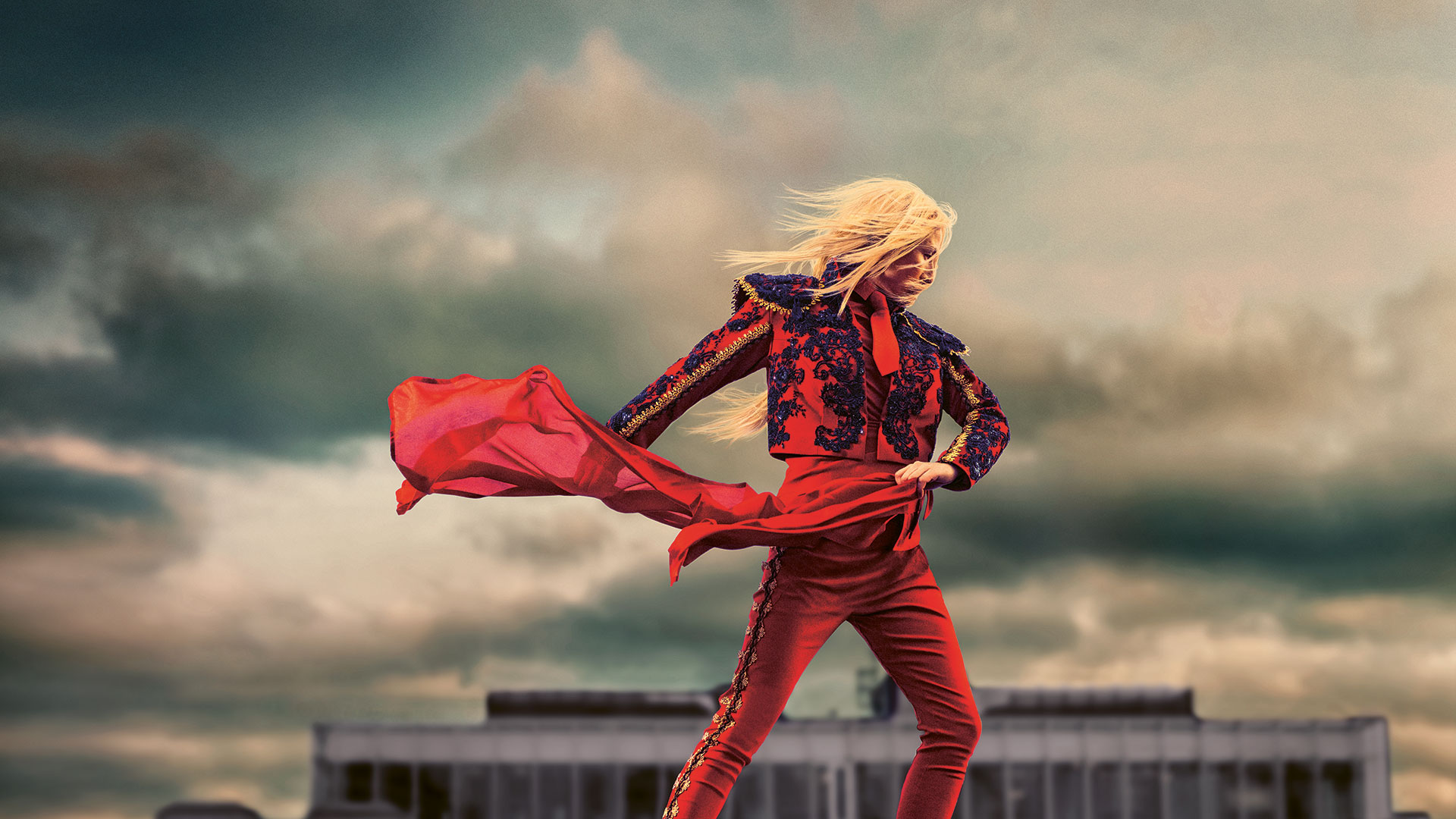 En figur i röd dräkt står framför en dramatisk himmel, med ett rött tyg som dansar i vinden och siluett av ett hus i bakgrunden.