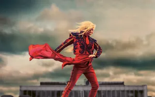 En figur i röd dräkt står framför en dramatisk himmel, med ett rött tyg som dansar i vinden och siluett av ett hus i bakgrunden.