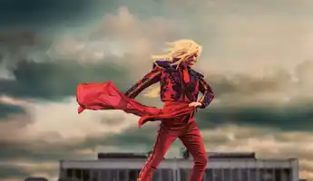 En figur i röd dräkt står framför en dramatisk himmel, med ett rött tyg som dansar i vinden och siluett av ett hus i bakgrunden.