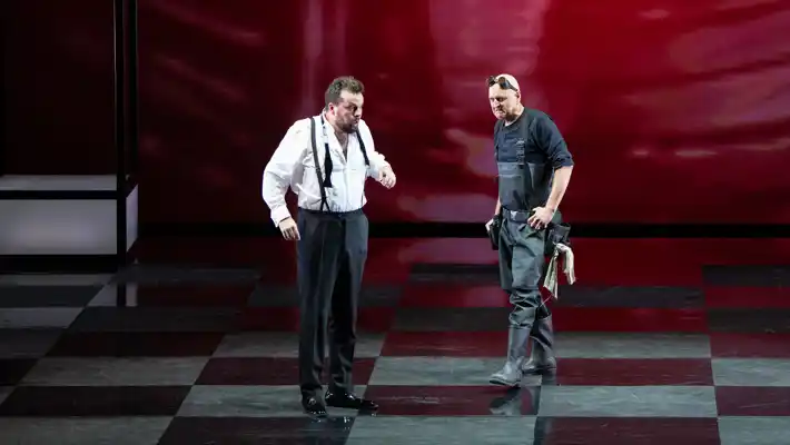 Två män står på en scen, golvet är rutigt, bakom dem en röd vägg.