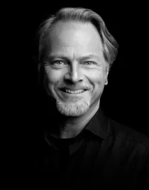 En närbild på en leende man med skägg i mörk skjorta.