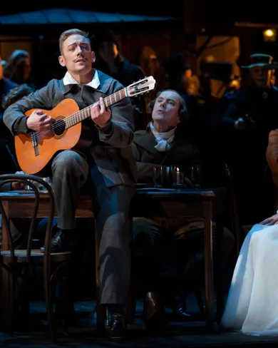 A group of people sitting around a table with a man playing the guitar.