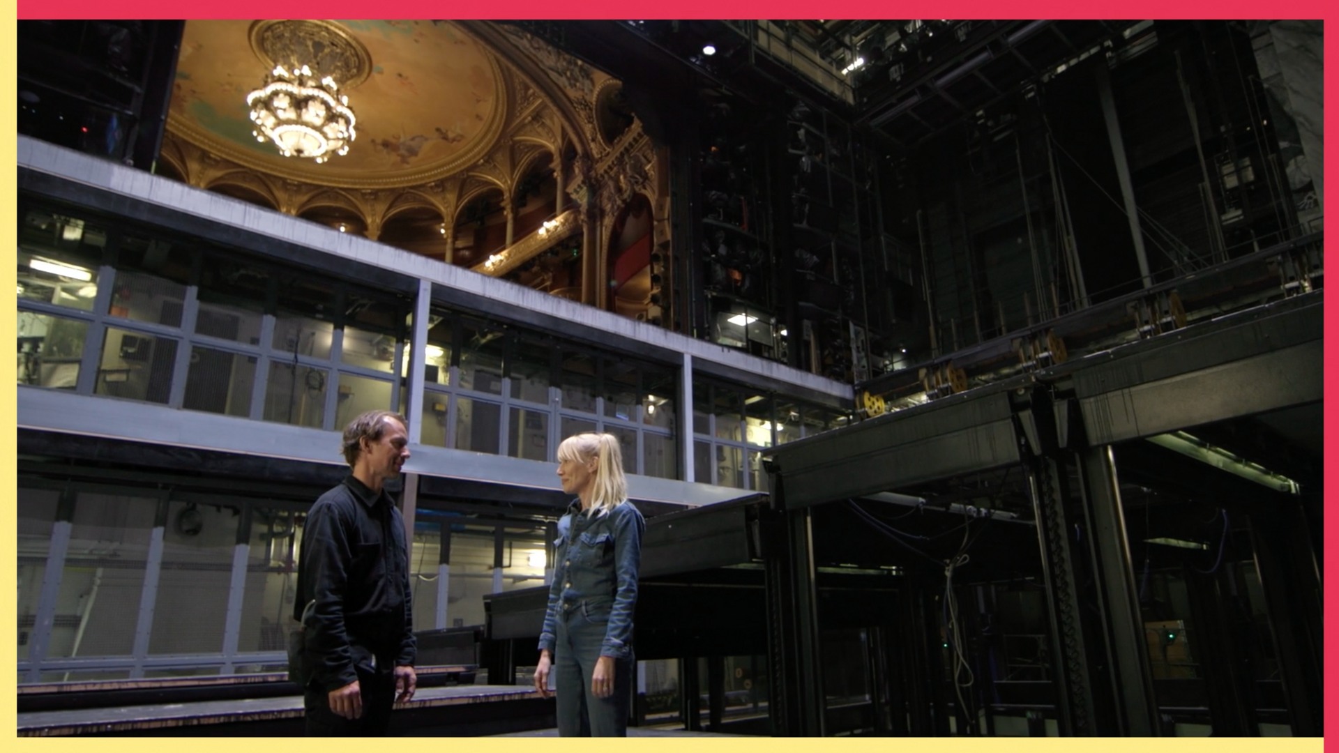 En man och en kvinna står i ett stort scenbygge, i bakgrunden skymtar en publiksalong.