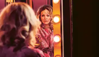 A woman sits with her back to us, she looks into a mirror surrounded by lights.