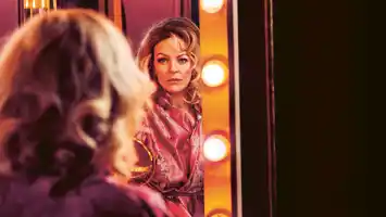 A woman sits with her back to us, she looks into a mirror surrounded by lights.