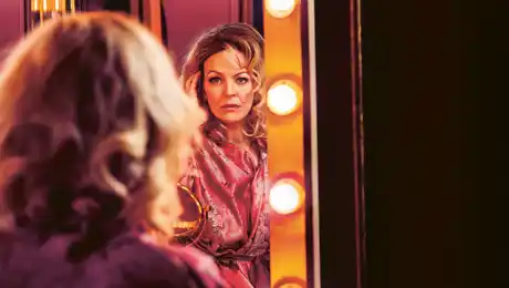 A woman sits with her back to us, she looks into a mirror surrounded by lights.
