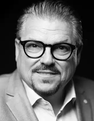 Närbild på en man med glasögon: han bär en kavaj och uppknäppt skjorta.