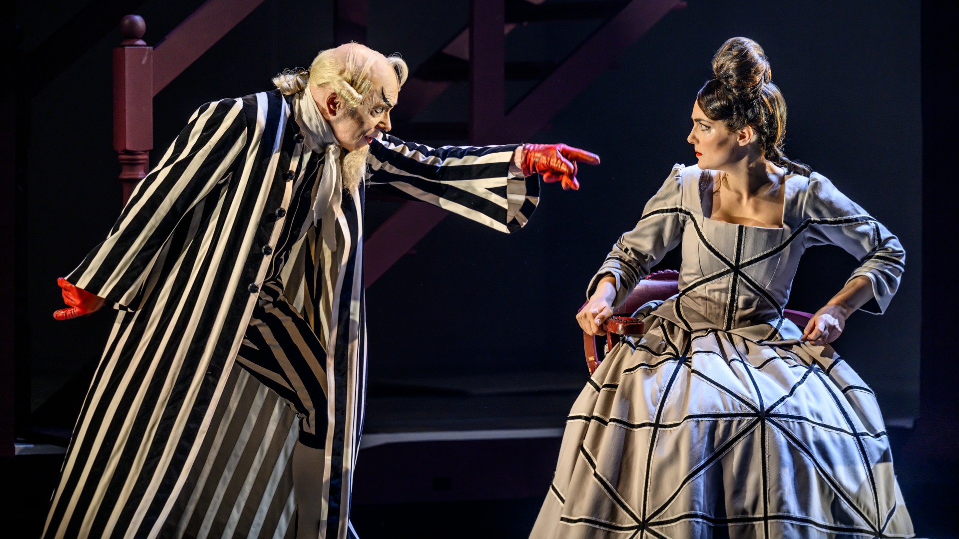 En man i randig kostym och röda handskar pekar på en kvinna i rutig klänning.