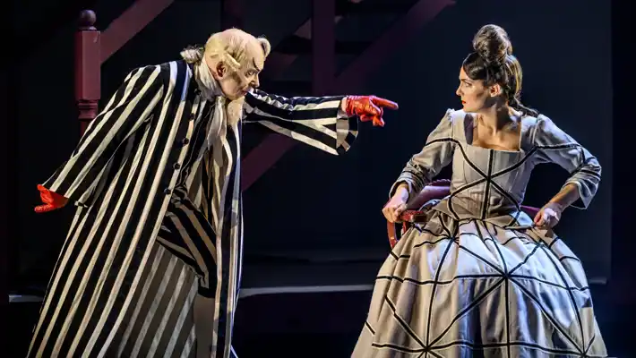 A man in a striped suit and red gloves points at a woman in a checkered dress.