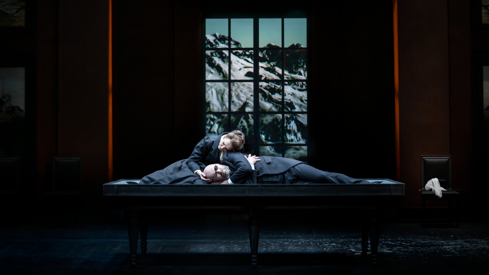 A woman and a man are half-reclining on a large table, in the background a window overlooking an Alpine landscape.