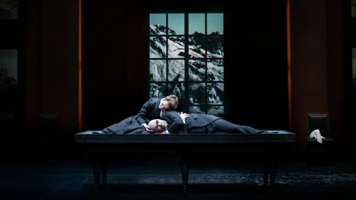 A woman and a man are half-reclining on a large table, in the background a window overlooking an Alpine landscape.