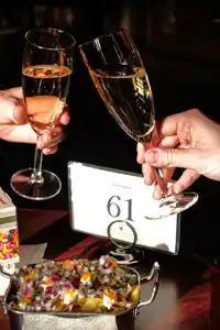 Two people toast with champagne in glasses, various snacks are on the table.