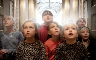 Grupp av barn i olika kläder står tillsammans, inomhus med guldkolumner i bakgrunden.